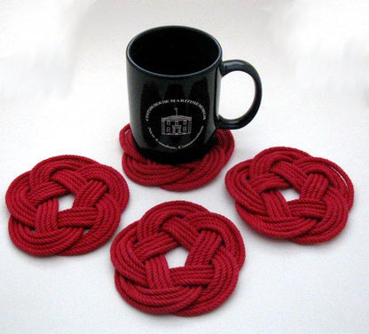 A black mug rests on a red Nautical Sailor Knot Coaster from Mystic Knotwork, handcrafted from cotton, with three more coasters from the set of four arranged nearby on a white surface—each one proudly handmade in the USA.