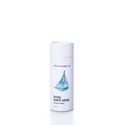 A white cylindrical container labeled “Shore Soap Co. Smooth Sailing Eco-Friendly Body Balm Stick,” featuring an illustration of a sailboat on the front, stands upright against a white background, highlighting this pocket-sized moisturizer for on-the-go hydration.
