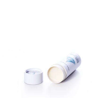 A cylindrical Smooth Sailing Eco-Friendly Body Balm Stick by Shore Soap Co, with its lid removed and lying on its side, reveals the solid pocket-sized moisturizer inside against a white background.