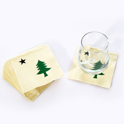 A glass rests on a Maine Bicentennial Cocktail Napkin by Boston International, decorated with a green Christmas tree and black star reminiscent of the pine-tree-and-star flag, while a stack of identical napkins sits nearby. by Sea Bags