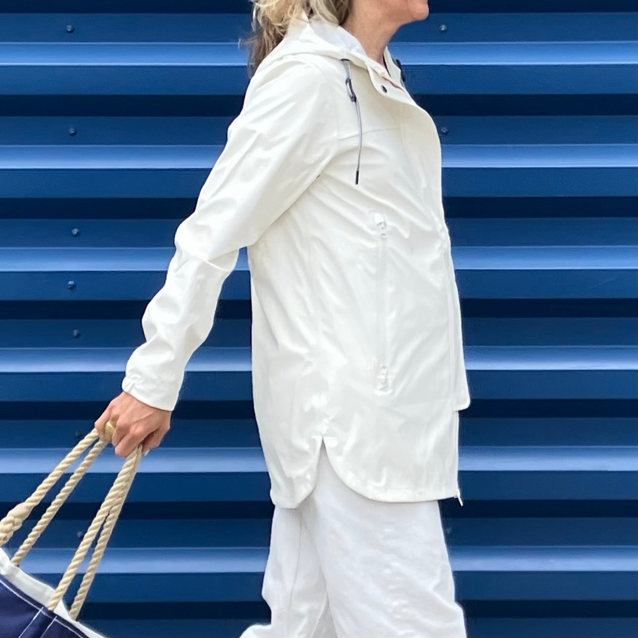 Wearing the Harborside Waterproof Raincoat in white by Batela USA Inc, a person walks while pulling a navy bag with rope handles in front of a blue corrugated metal wall, ready for any coastal weather.