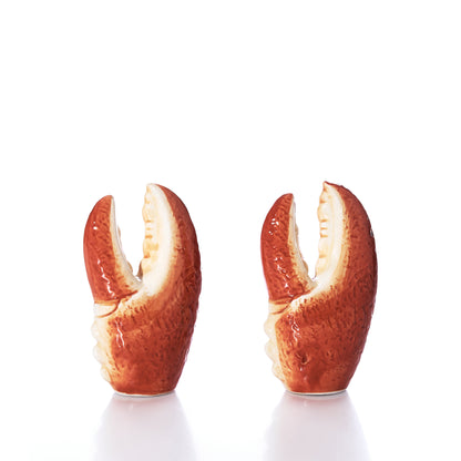 The Lobster Claw Salt  Pepper Shaker Set by Batela USA Inc features two upright ceramic shakers shaped like lobster claws, displayed on a white surface with a white background. These charming shakers bring unique seaside flair to your kitchen décor.