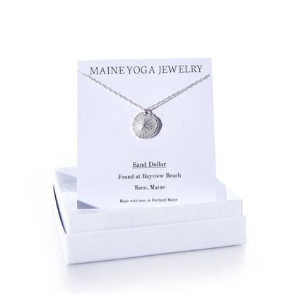 A Pendant Necklace - Sand Dollar Silver by Maine Yoga Jewelry rests on a card inside a white box. The card notes that the sand dollar was found at Bayview Beach, Saco, Maine, emphasizing the brands sustainable craftsmanship inspired by Maines natural beauty. by Sea Bags