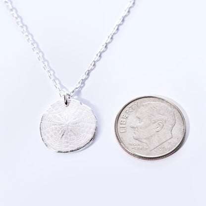 The Pendant Necklace - Sand Dollar Silver by Maine Yoga Jewelry features a round, textured silver sand dollar charm—handcrafted in Maine with sustainable craftsmanship—and is pictured next to a U.S. dime on a white background for size reference. by Sea Bags
