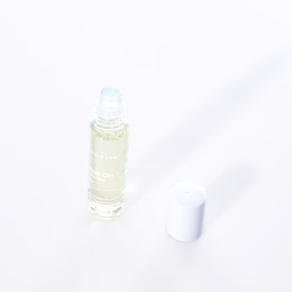A clear glass roll-on bottle of Shore Soap Cos Warm Sand Eco-Friendly Roll-On Fragrance, featuring an alcohol-free jasmine scent, with a white plastic cap placed beside it on a white background. by Sea Bags