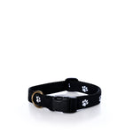 The Belted Cow Paw Print Dog Collar 1″ - Medium is crafted from durable black nylon webbing, features a plastic buckle, sturdy metal ring, and is decorated with a white paw print pattern, shown on a white surface.