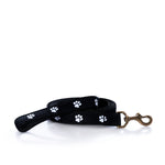 The Belted Cow Paw Print Dog Leash, featuring a black design with white paw prints and a robust metal clip, is coiled neatly on a white background.