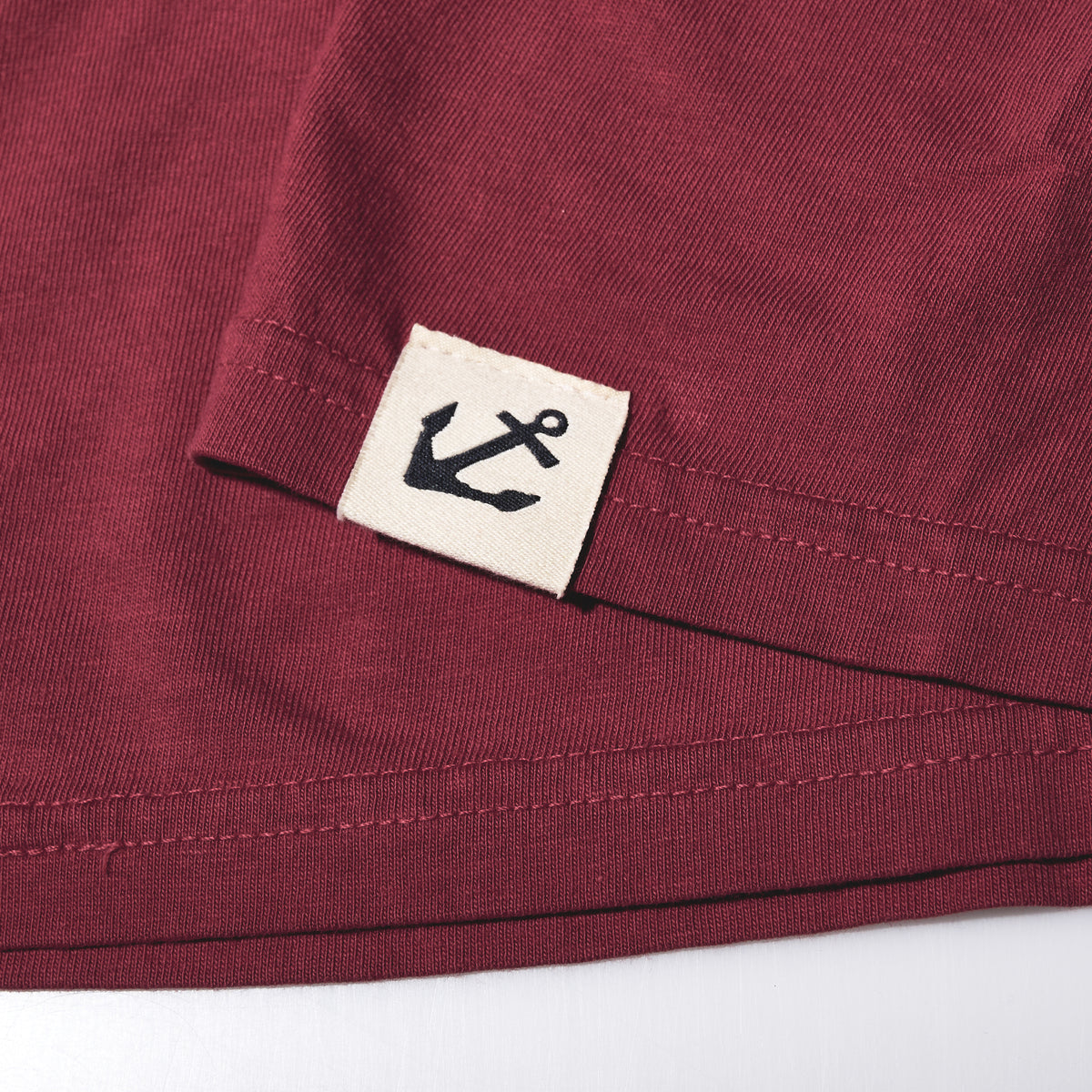 Close-up of a maroon East Coast Shark T-Shirt with a small square label on the hem featuring a black anchor icon.