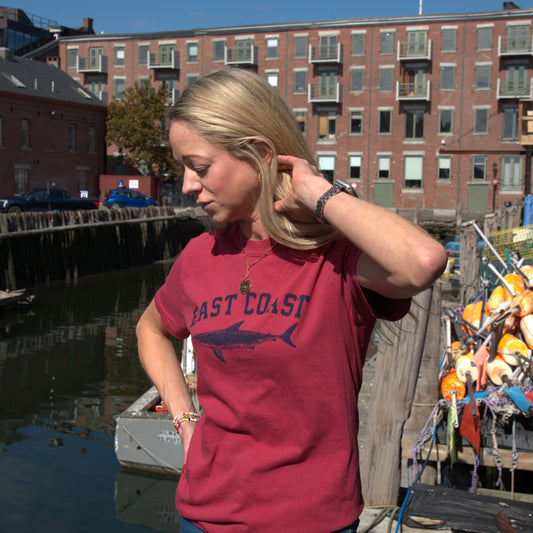 A woman wearing a maroon East Coast Shark T-Shirt with a shark silhouette stands on a wooden dock by a harbor.