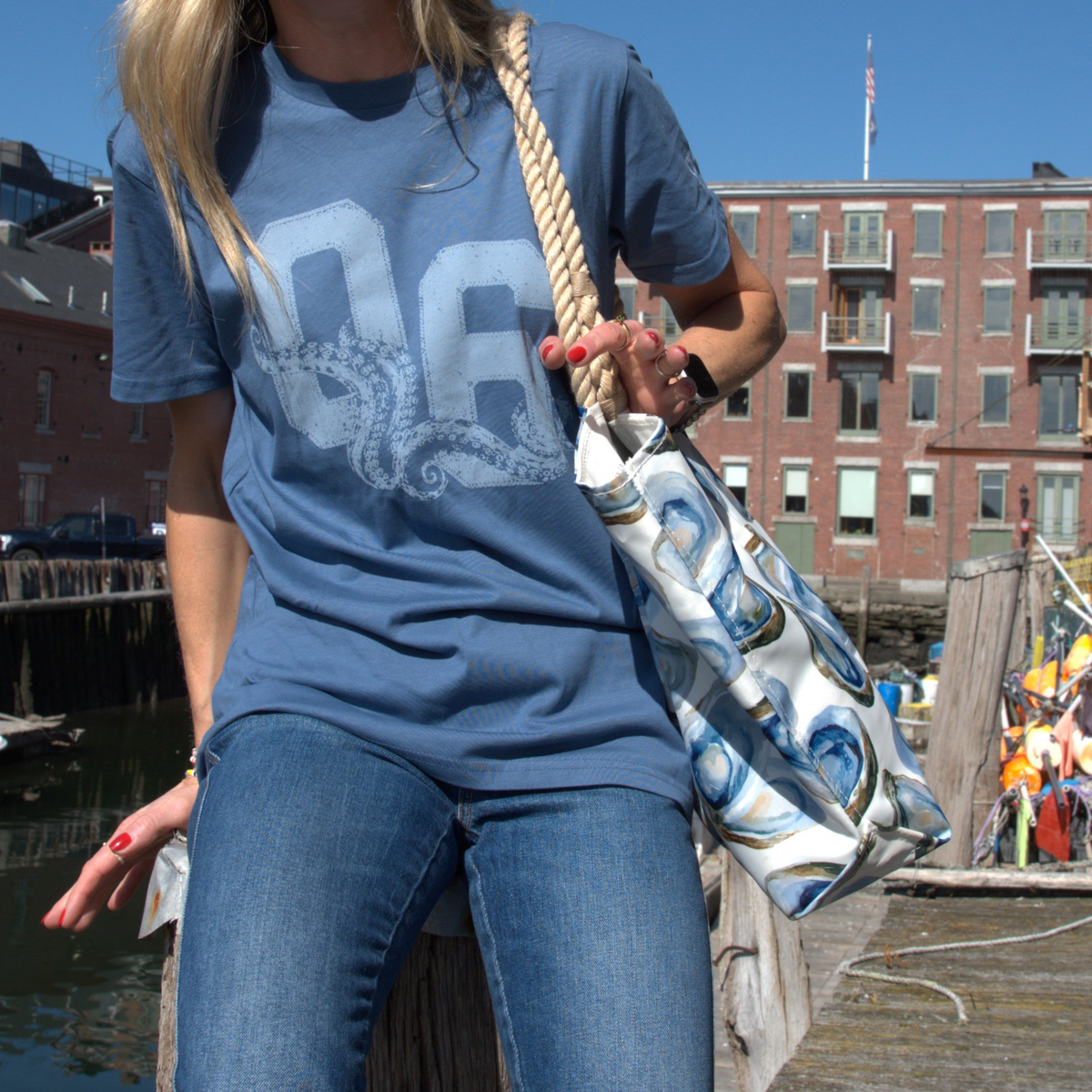 A woman wearing a blue graphic t-shirt and jeans stands on a wooden dock carrying a white tote bag with a blue and white oyster pattern and a thick rope handle, with a harbor and brick buildings in the background.