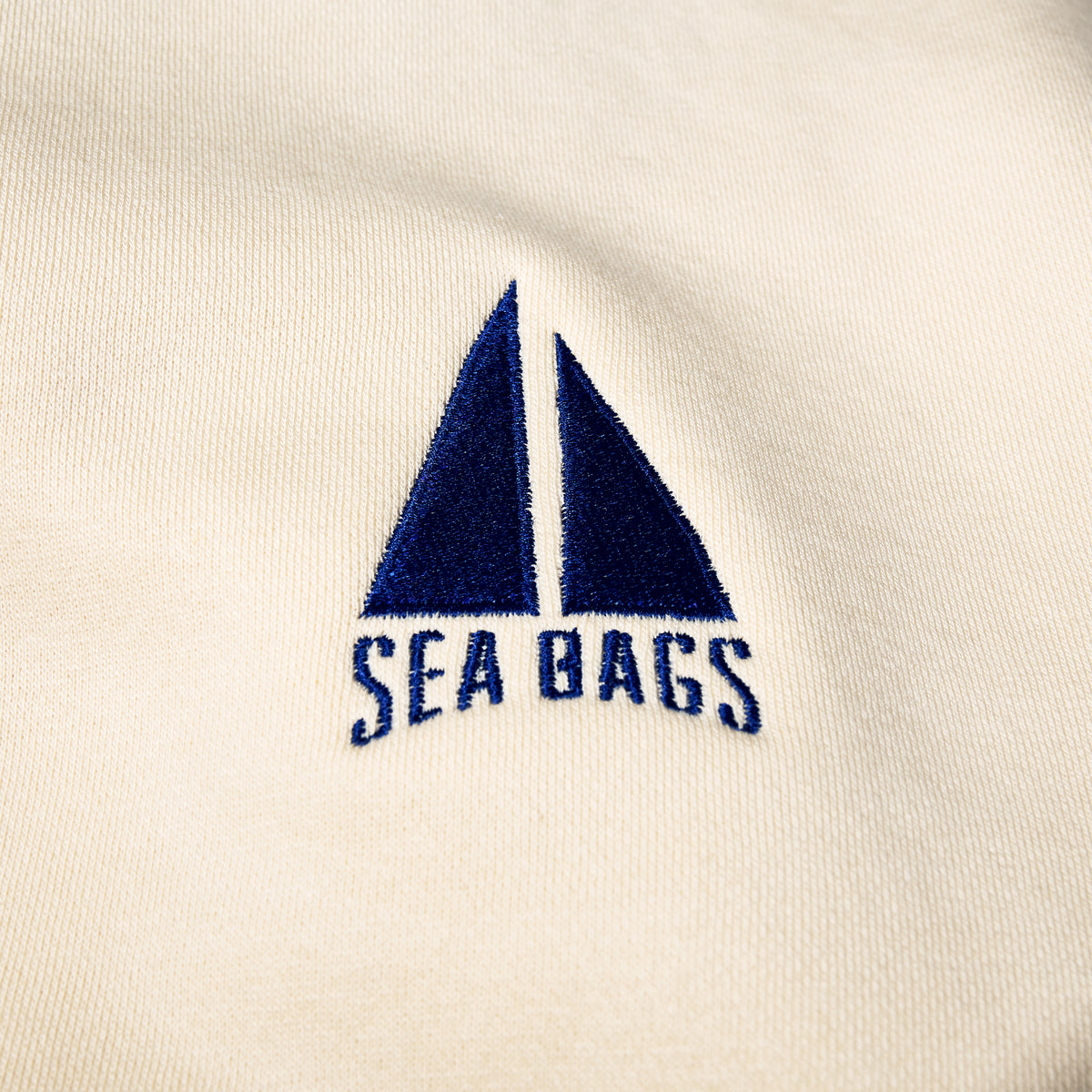 Cream-colored Sailboat Crewneck Sweatshirt featuring an embroidered dark blue sailboat icon above the text “SEA BAGS.”
