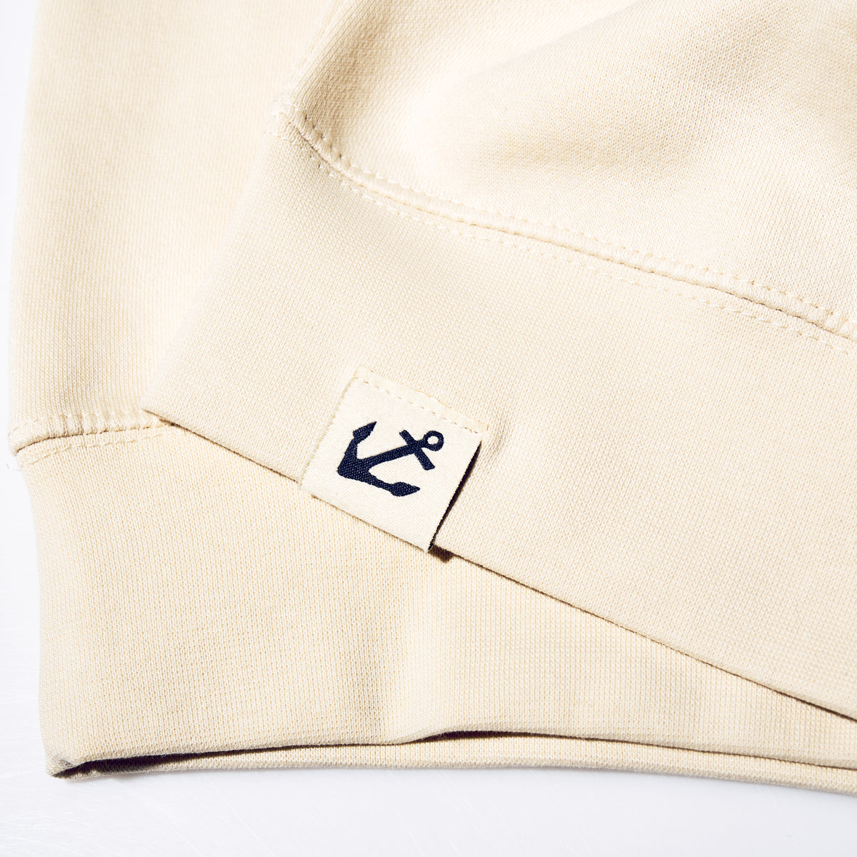 Close-up of a beige cotton sleeve hem on a Sailboat Crewneck Sweatshirt featuring a small square tag with a navy blue anchor icon.