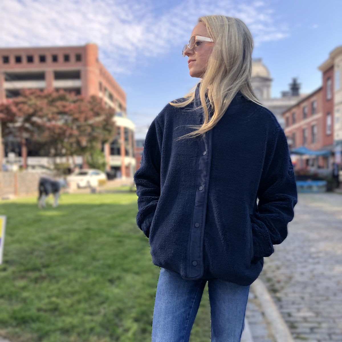 A person wearing a navy Fleece Snap-Front Jacket - Navy over blue jeans, standing in a city square.