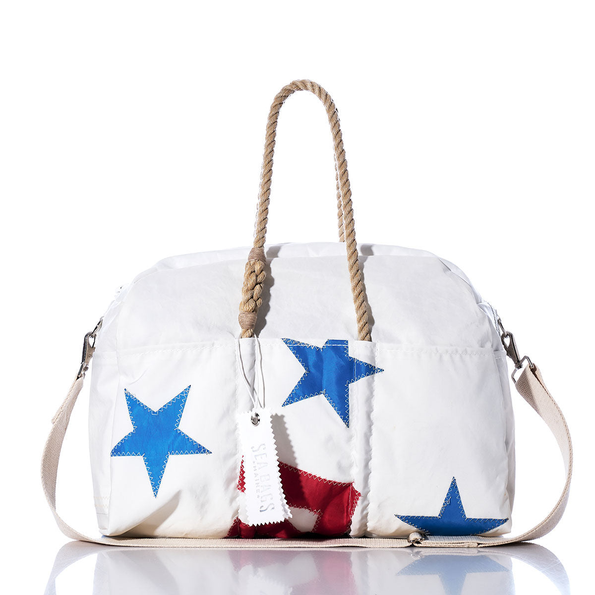 Meet the Vintage Ensign Weekender by Sea Bags—a white weekender bag featuring bold blue stars, a vibrant red pattern, tan rope handles, and a beige shoulder strap. Shown on a white background, this stylish travel essential is your perfect go-to for vintage-inspired getaways. by Sea Bags