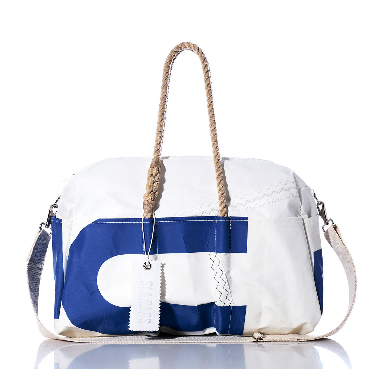 The Sea Bags Vintage US Weekender—a white and blue bag featuring rope handles, a fabric shoulder strap, and a small white tag attached to the handle—is photographed on a reflective surface. by Sea Bags