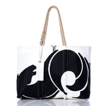 The Vintage Beneteau Large Tote by Sea Bags features a bold black abstract swirl on white sailcloth, finished with rope handles and a white tag. Photographed against a white background, this eye-catching tote blends coastal charm with the renowned durability of handmade Maine craftsmanship. by Sea Bags