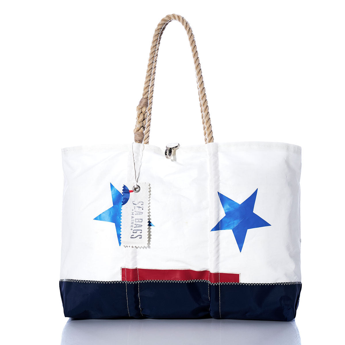 The Vintage Ensign Ogunquit Beach Tote by Sea Bags features a white recycled sail cloth design with blue stars, a red stripe, and a navy bottom. It has sturdy rope handles and a Sea Bags tag. The tote is photographed on a plain white background with a subtle reflection beneath it. by Sea Bags