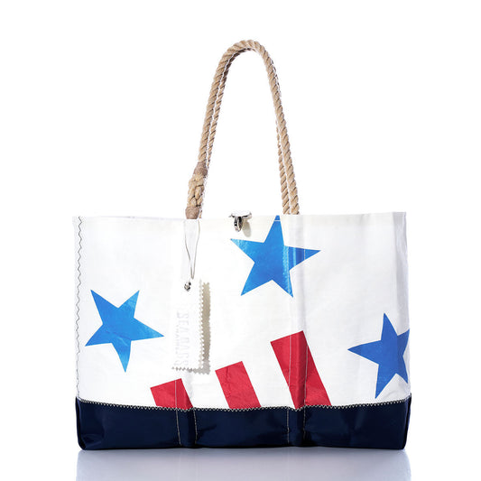 The Sea Bags Vintage Ensign Ogunquit Beach Tote is a large white beach bag handcrafted from recycled sail cloth, featuring a blue bottom, sturdy rope handles, and a design of blue stars and red stripes inspired by a vintage ensign. A white tag is attached to one handle. by Sea Bags