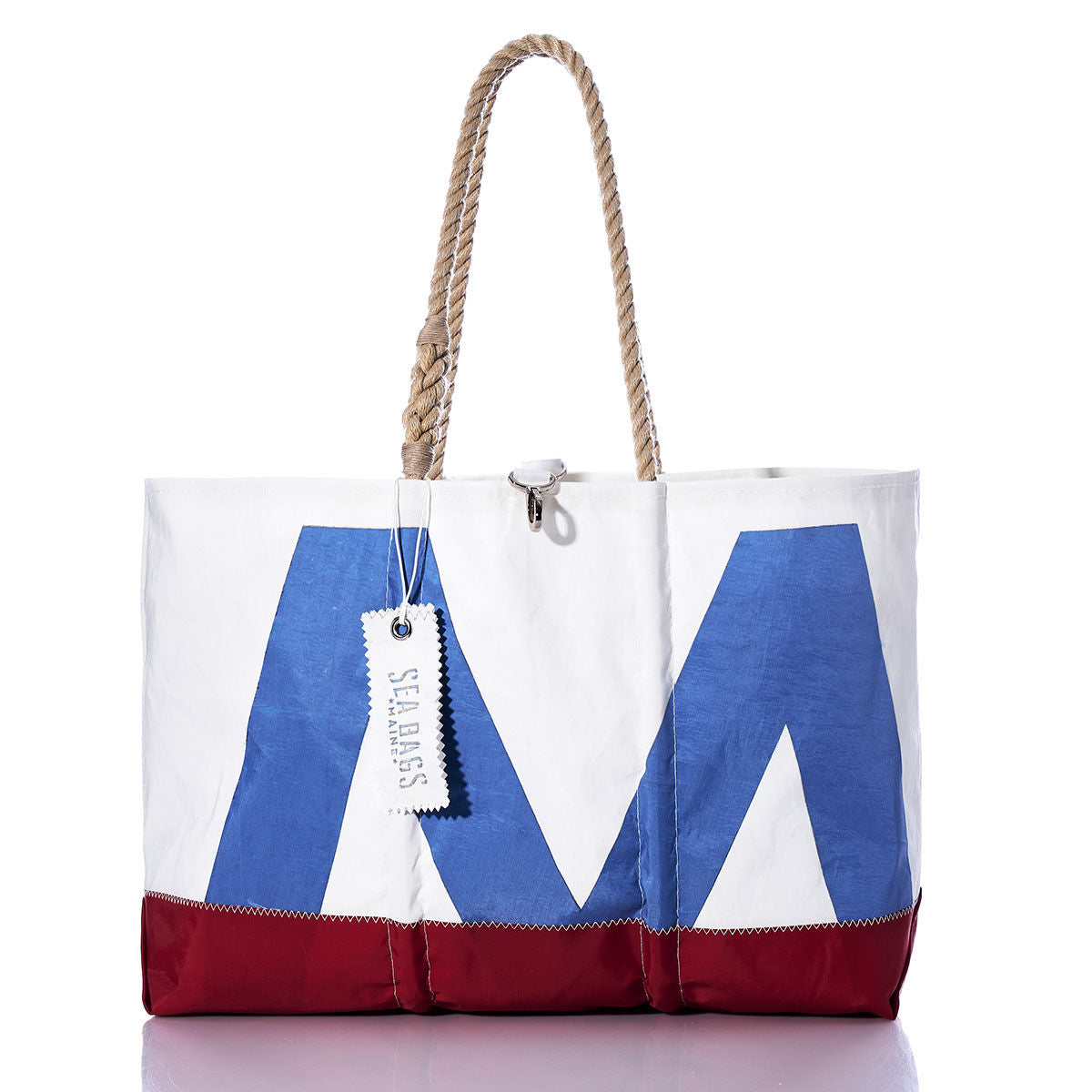 Crafted from recycled sail cloth, the Vintage Blue M Ogunquit Beach Tote by Sea Bags features a large blue M design on the front, accented with red and blue details. This white beach tote includes rope handles and an attached Sea Bags tag, combining coastal style with eco-friendly materials. by Sea Bags
