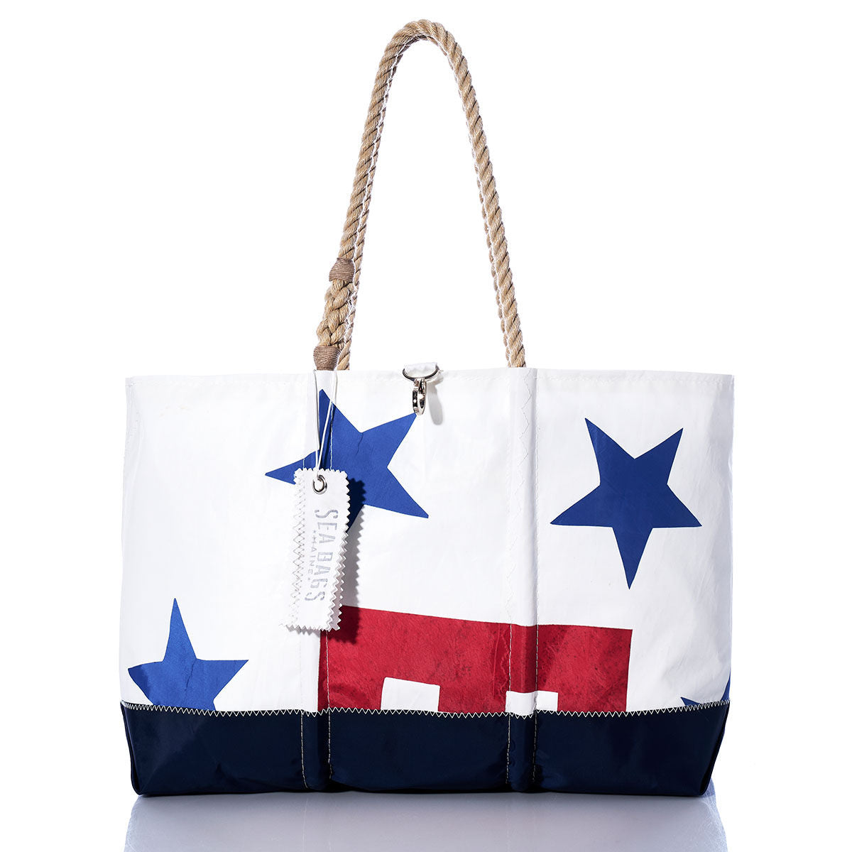 Introducing the Vintage Ensign Ogunquit Beach Tote from Sea Bags—a white tote bag crafted from recycled sail cloth, featuring blue stars, a striking red rectangle, and a navy base. Finished with rope handles and a hanging white tag with text, this stylish and eco-friendly accessory is perfect for your seaside adventures. by Sea Bags