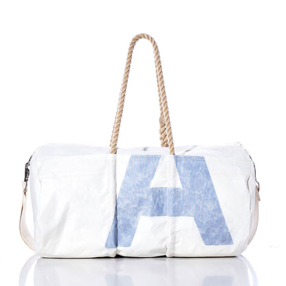 Introducing the Sea Bags Vintage Blue U Duffel—a white vintage duffel bag crafted from recycled sail cloth, featuring a bold blue letter U on the front. This stylish bag is finished with sturdy hemp rope handles and a beige adjustable shoulder strap, and is photographed against a crisp white background. by Sea Bags