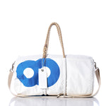 Introducing the Sea Bags Vintage 869 Duffel—this distinctive bag is crafted from recycled sail cloth and features a striking blue circular motif, complemented by sturdy hemp rope handles, a beige shoulder strap, and a signature white tag attached to the handle. The entire piece is beautifully showcased on a reflective surface. by Sea Bags