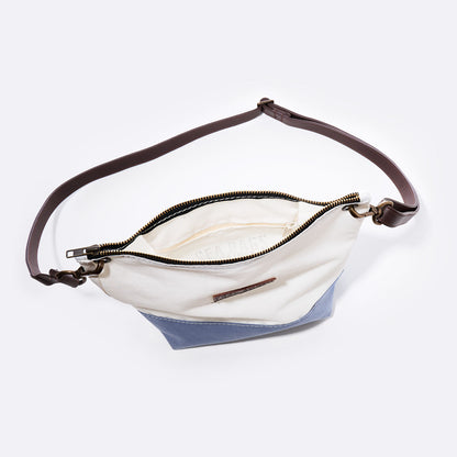 The Chambray Blue Chebeague Crossbody Bag by Sea Bags, crafted from recycled sail cloth, features a two-tone design with a brown adjustable strap, gold zipper, and white interior lining. Part of the Chebeague Collection, this nautical bag is displayed open against a plain background. by Sea Bags