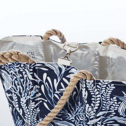 The Navy Ocean Flora Handbag by Sea Bags—a navy blue tote adorned with white leaf patterns, beige rope handles, and a silver clip closure—crafted from recycled sail cloth for an earth-friendly nod to coastal life, is shown here against a white background. by Sea Bags