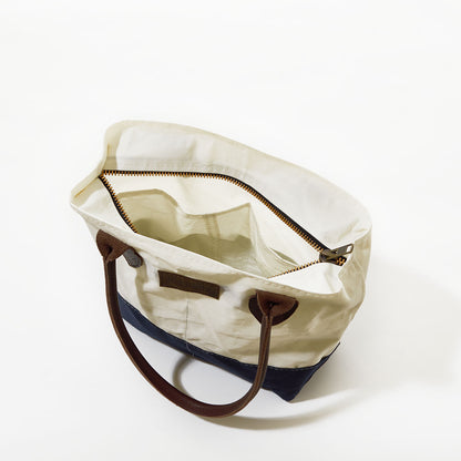 The Sea Bags Chebeague Handbag from the Chebeague Collection is an eco-friendly nautical tote crafted from recycled sail cloth, featuring brown handles, a white upper section, a dark blue bottom, and a visible zippered inner pocket. The bag is photographed from above on a white background. by Sea Bags
