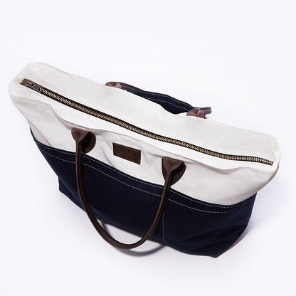 The Sea Bags Chebeague Large Travel Tote is crafted from water-resistant canvas, featuring a stylish white top, navy bottom, brown leather handles, and a top zipper—making it the perfect carry-on bag for travel. by Sea Bags