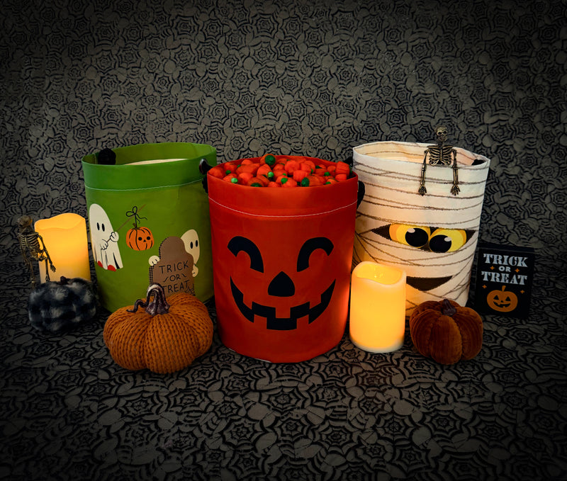 A Halloween-themed product display featuring three decorative bucket costumes: a green bucket with a ghost and pumpkin design, a red bucket with a jack-o'-lantern face, and a white bucket wrapped in faux bandages with spider details. The buckets are styled with small knit pumpkins, glowing candles, and a 'Trick or Treat' sign against a dark patterned background.