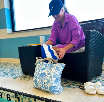 A person in a purple shirt and black cap sits by an indoor pool, reaching into the Sea Bags Paisley Mermaid Medium Tote, with a striped towel nearby. White sandals are on the floor. by Sea Bags