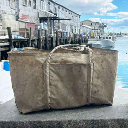 A weathered Sea Bags canvas Jeb tote bag with rope handles rests on a concrete surface in front of a waterfront dock, surrounded by boats and old buildings.