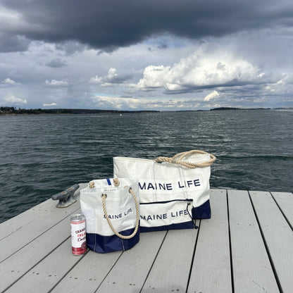 Two Sea Bags Maine Life Large Totes and a can of cranberry lime seltzer rest on a dock by a large body of water under a cloudy sky, capturing the spirit celebrated by Erin Ovalle. by Sea Bags