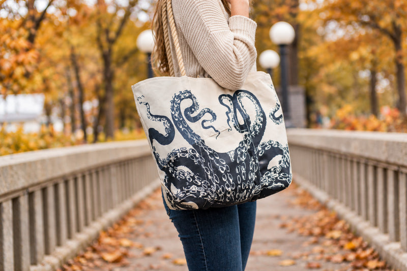 A person outdoors on a bridge carrying a large cream canvas tote bag with a dark navy octopus tentacle print. The background shows blurred autumn trees with orange and yellow leaves.