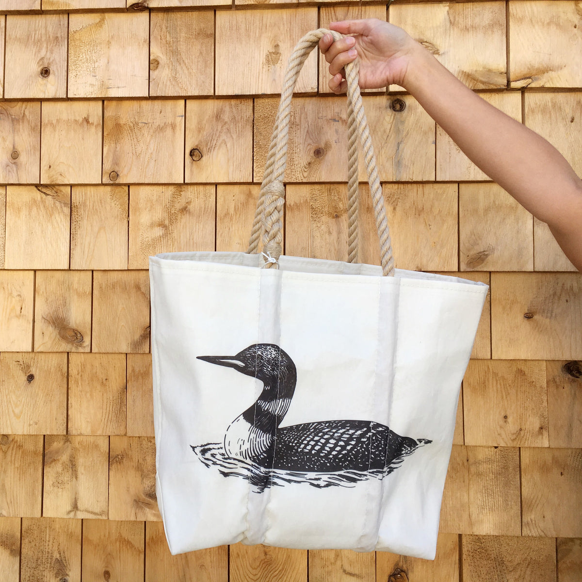 A hand holds a white recycled sail cloth Loon Medium Tote by Sea Bags, featuring thick rope handles and a black graphic of a Maine loon swimming on water.