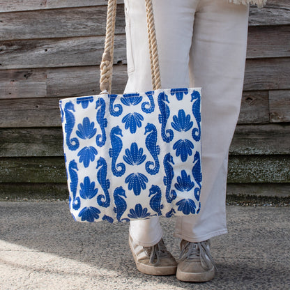 Person on pavement holding a Sea Bags Blue Sea Horse Print Handbag, a white tote with blue seahorse and shell print and thick rope handles.