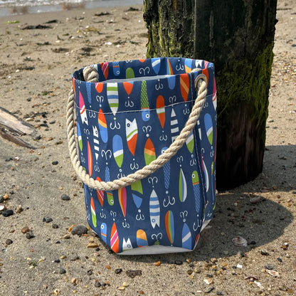 The Kate Nelligan Lures Bucket Bag by Sea Bags, featuring a vibrant fish-patterned coastal print and sturdy rope handle, sits on the sand near a wooden post at the beach—making it the perfect nautical essential for seaside adventures. by Sea Bags