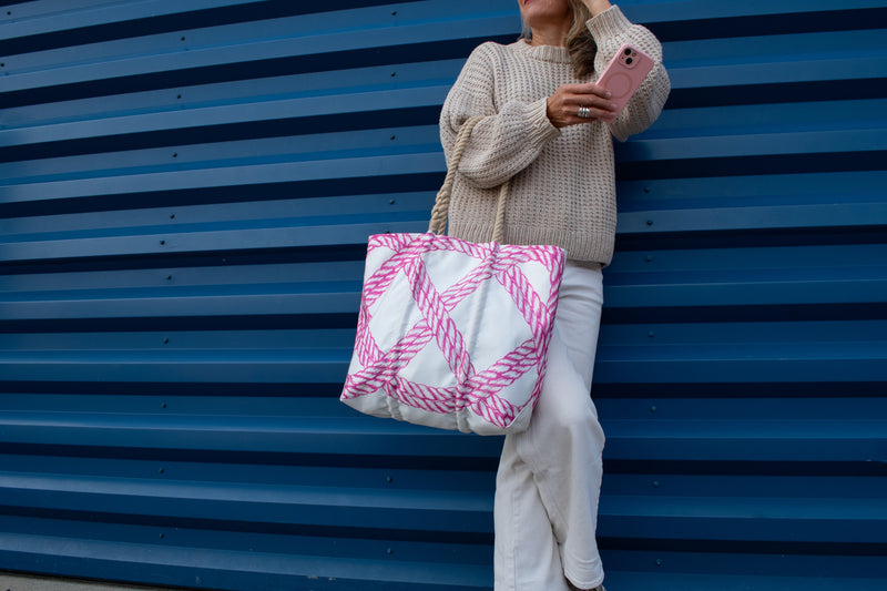 A woman wearing a beige knit sweater and white pants stands against a blue corrugated metal wall, holding a large white tote bag with pink rope-style woven accents.