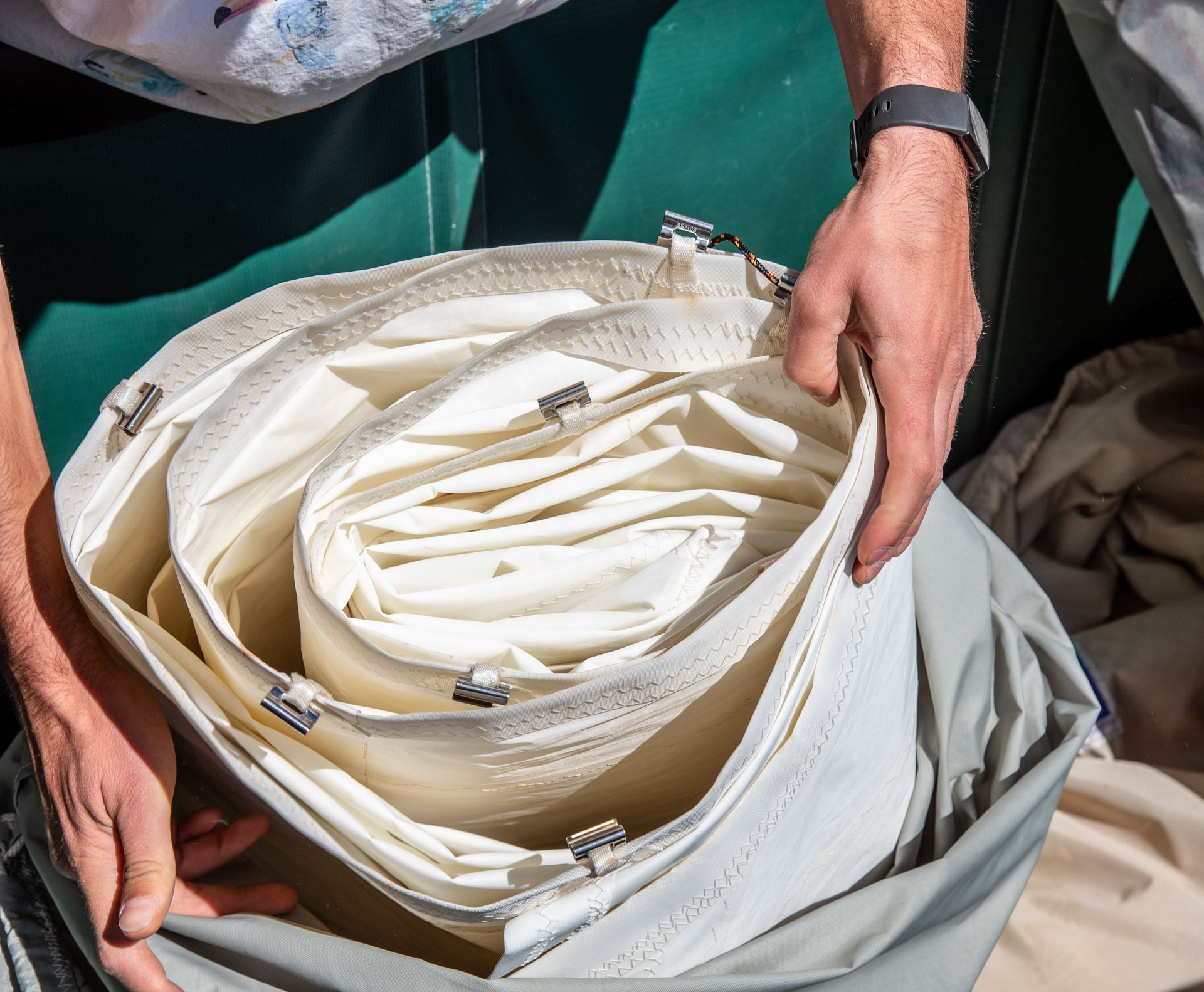 A person with a black wristwatch is holding a coiled, white, fabric-like material secured with metal clasps -similar to what’s used in crafting sturdy sail bags or stylish Sea Bags tote bags