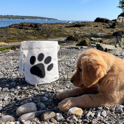 A golden retriever puppy lies on a rocky beach beside the Sea Bags Paw Print Bucket Bag, a white storage bucket featuring a black paw print, with water and trees in the background. by Sea Bags