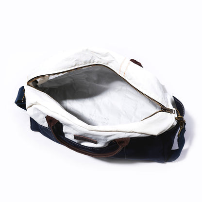 Top view of the Chebeague Weekender by Sea Bags, featuring white lining, brown leather handles, a blue body made from recycled sail cloth, and a brass zipper. The empty bag rests on a white background. by Sea Bags