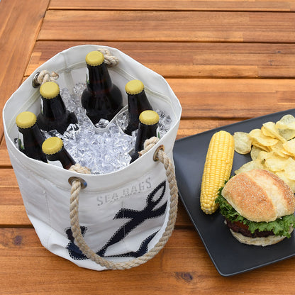 A Four Leaf Clover Beverage Bucket Bag by Sea Bags, crafted from recycled sail cloth, in white and collapsible design, sits filled with ice and six bottled drinks next to a plate holding a hamburger, corn on the cob, and potato chips on a wooden table. by Sea Bags