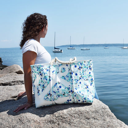 A person sits on a rocky shore holding a White Anchor on Sea Glass Print Large Tote, which features a white anchor pattern against a blue sea glass mosaic print.