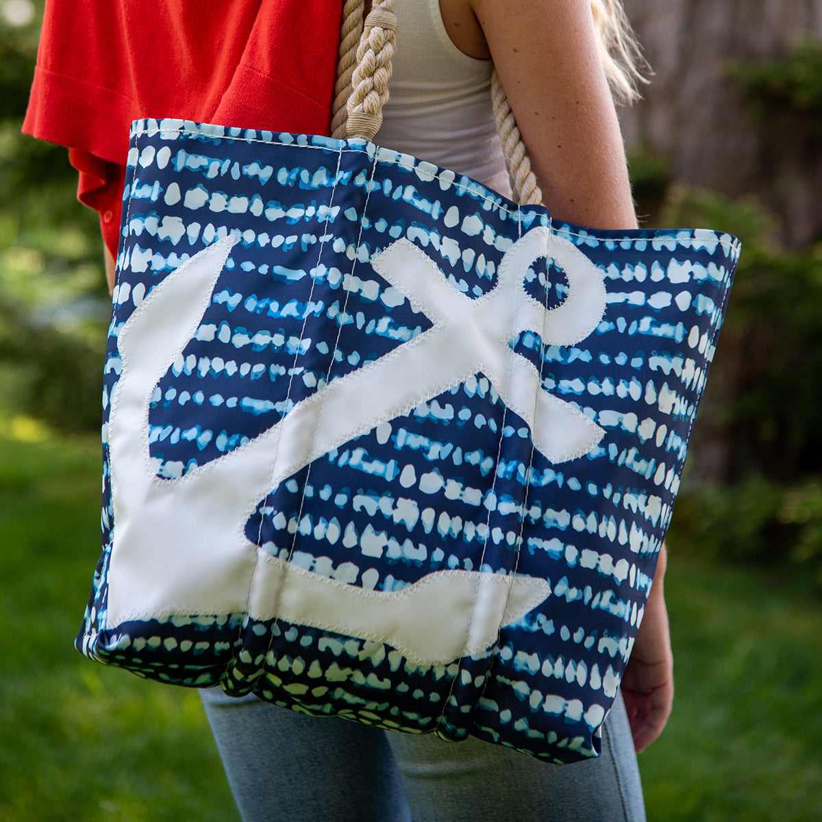 A person carries the Sea Bags White Anchor on Shibori Print Tote, featuring a bold anchor design and sturdy rope handles, made from recycled sail cloth. Grass and trees are visible in the background. by Sea Bags