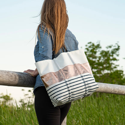 A woman with long hair stands by a wooden fence, carrying the Sea Bags Zip Top Rose Gold-on-Navy Mariner Stripe Medium Tote. Green grass and trees are in the background. by Sea Bags