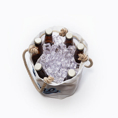 Six brown glass bottles in a white Sea Bags UMaine Beverage Bucket filled with ice cubes, shown from above. The bucket, crafted from recycled sail cloth, features thick rope handles and blue lettering on the side—evoking the classic style of a University of Maine bucket. by Sea Bags