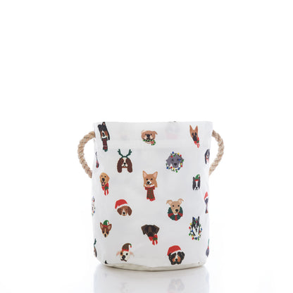 The Sea Bags Very Merry Pups Bucket Bag is a white fabric storage basket featuring rope handles and playful cartoon dog faces adorned with festive accessories like hats, scarves, reindeer antlers, and wreaths—an eco-friendly gift basket that’s perfect for any dog lover and a delightful touch to your holiday décor. by Sea Bags