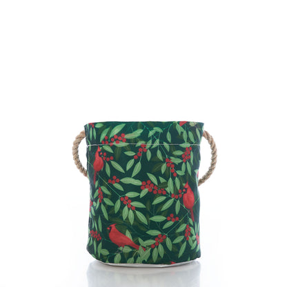 The Sea Bags Winterberry Cardinals Bucket Bag, crafted from sustainable fabric and featuring rope handles, is beautifully adorned with a cardinal and red berries motif on a green leafy background. Shown against a crisp white surface and backdrop, this eco-friendly bag adds festive charm to your holiday decor. by Sea Bags