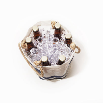 Six amber glass bottles of beer, chilled in ice, are arranged inside the Sea Bags Breton Stripe Whale Beverage Bucket—a white recycled sail cloth cooler with thick rope handles—viewed from directly above against a white background. by Sea Bags